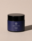 A 50g jar of Divinum Celeste Crème cosmetic cream, featuring a deep purple label and a black lid, set against a warm beige background with a soft shadow.