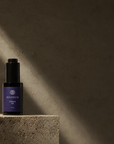 A small violet bottle with a dark purple label reading Divinum Halcyon Oil, placed on a textured stone pedestal in dramatic side lighting against a dark stone wall.