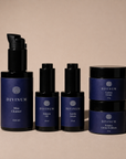Five Divinum skincare bottles and jars arranged on a warm beige studio background. The set includes a pump bottle of Mira Cleanser, two small bottles of Halcyon Oil and Aurelia Serum, and two stacked jars of Celeste Creme and Solstice Creme Exfoliant.