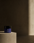 A small violet glass cosmetic jar with a black lid sits on a textured beige pedestal in a dimly lit studio setting with a warm spotlight on the right.