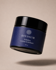A floating cosmetic jar of Divinum Solstice Crème Exfoliant, featuring a deep purple label and a black lid, set against a warm beige background with a soft shadow below.