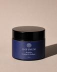 A 50g jar of Divinum Solstice Crème Exfoliant, featuring a deep purple label and a black lid, set against a warm beige background with a soft shadow.