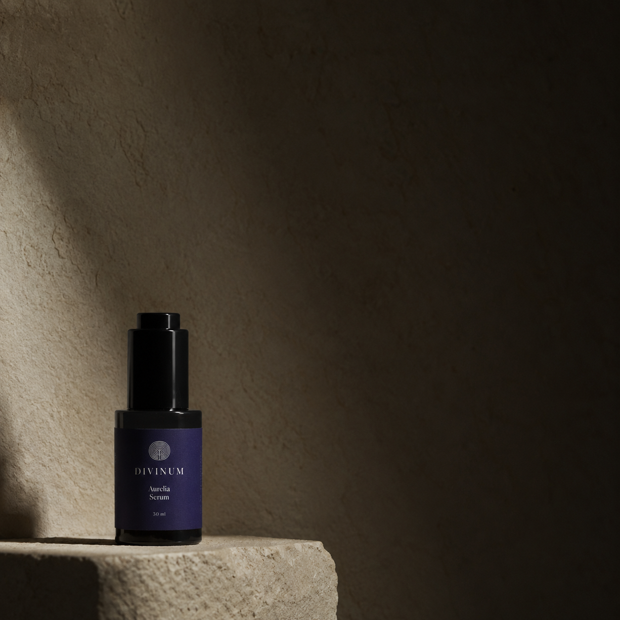 A dark violet bottle with a purple label and black cap, standing on a textured stone pedestal against a dark beige wall. The label reads Divinum Aurelia Serum.