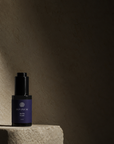 A dark violet bottle with a purple label and black cap, standing on a textured stone pedestal against a dark beige wall. The label reads Divinum Aurelia Serum.