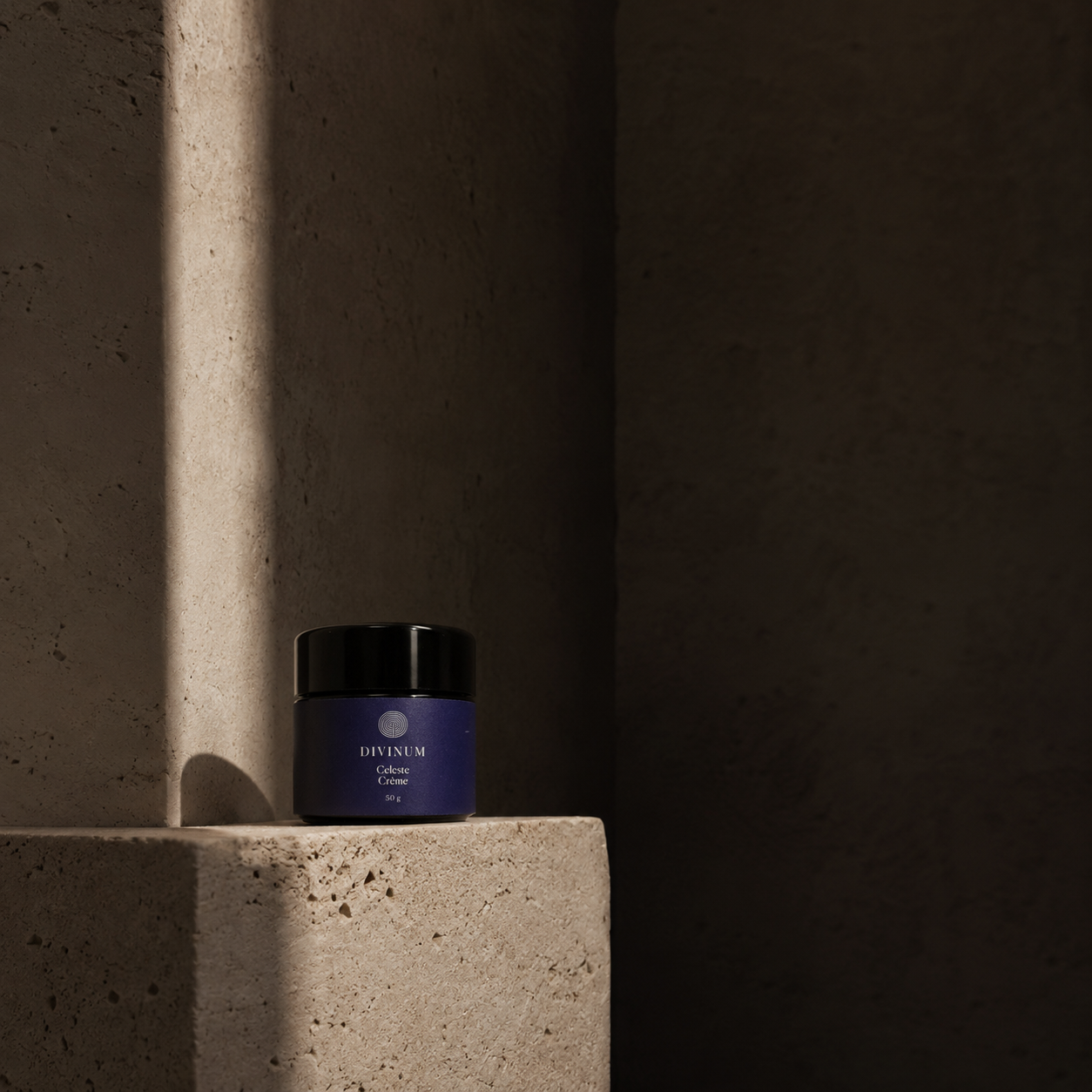 A small jar of Divinum Celeste Creme skincare product with a dark purple label and black lid, placed on a textured stone pedestal in dramatic side lighting against a dark concrete background.