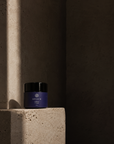 A small jar of Divinum Celeste Creme skincare product with a dark purple label and black lid, placed on a textured stone pedestal in dramatic side lighting against a dark concrete background.