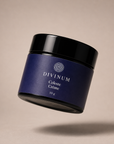 A floating cosmetic jar of Divinum Celeste Crème 50g, featuring a deep purple label and a black lid, set against a solid beige background with a soft shadow below.