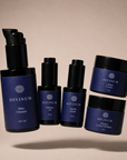 Five Divinum skincare products arranged on a warm beige studio background: a large violet pump bottle labeled Mira Cleanser, two smaller bottles labeled Halcyon Oil and Aurelia Serum, and two jars labelled Celeste Creme and Solstice Creme Exfoliant.