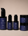 A set of five Divinum skincare products in dark violet and black packaging, arranged neatly on a warm beige background. The set includes a Mira Cleanser pump bottle, a Halcyon Oil dropper bottle, an Aurelia Serum dropper bottle, and two Creme jars stacked vertically.