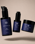 Three Divinum skincare products floating against a warm beige background: a large Mira Cleanser pump bottle, a smaller Halcyon Oil dropper bottle, and a Celeste Creme jar, all in dark violet and black packaging.