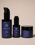 Three Divinum skincare products arranged against a solid beige background: a large black pump bottle of Mira Cleanser, a black dropper bottle of Halcyon Oil, and a black-lidded jar of Celeste Crème.